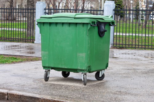 A large green plastic wheelie bin with a closed lid and four small wheels positioned on a paved surface, likely a driveway or sidewalk. The bin appears to be used for rubbish collection, with a slightly textured surface and a black handle on the lid. It is situated outdoors in front of a black metal fence with vertical bars and concrete posts, behind which there are trees and grass patches, suggesting a residential or public park area. The paved ground is slightly damp, indicating recent rain, and the environment is well-lit with natural daylight. This scene reflects typical rubbish storage in a community area that might be serviced by commercial waste removal companies such as Commercial Waste Marlow, servicing Marlow and surrounding areas postcode area.
