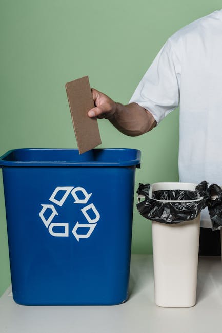 A person wearing a white shirt is shown holding a brown cardboard item in their right hand, positioned above a large blue recycling bin with a white recycling symbol printed on its front. The blue bin