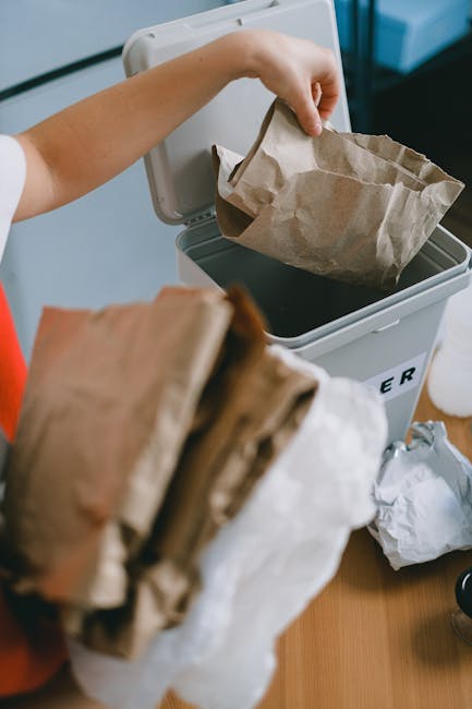 A person wearing a white shirt is shown holding a brown cardboard item in their right hand, positioned above a large blue recycling bin with a white recycling symbol printed on its front. The blue bin